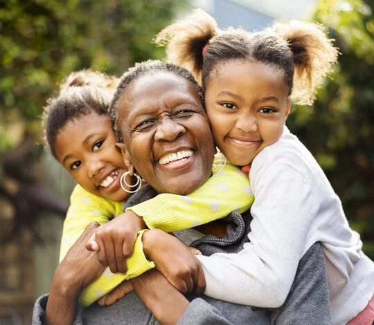 senior woman seated outdoors with her two grandchildren, who are embracing her from behind and smiling