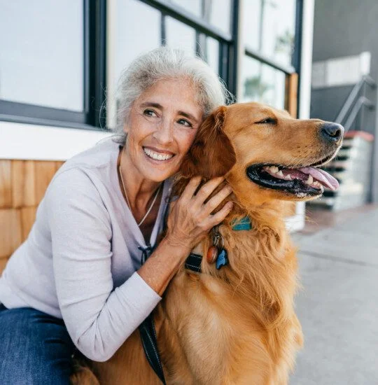senior woman smiles and kneels down to pet her smiling dog