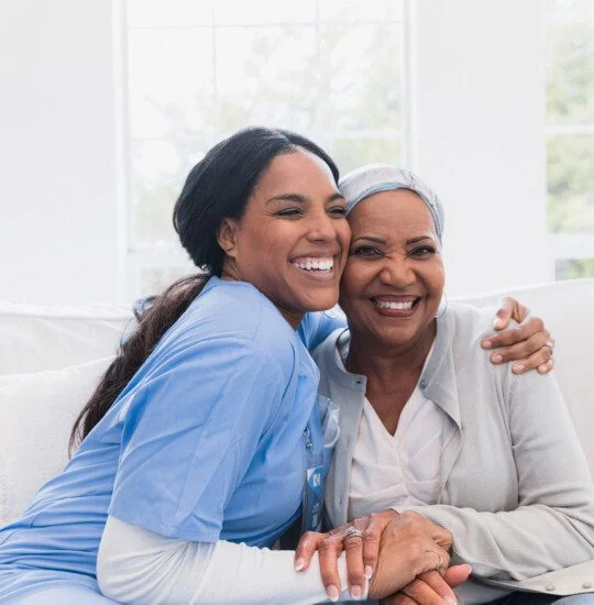 senior woman smiles and hugs her caregiver while seated on a couch in her apartment