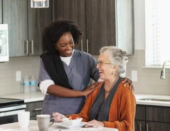 A home healthcare worker or nutritionist, an African-American woman in her 30s, helping a senior woman in her 80s eating a healthy snack in the kitchen. They are conversing and the senior is laughing. senior woman seated at the island in her home smiles while enjoying breakfast in the company of her caregiver