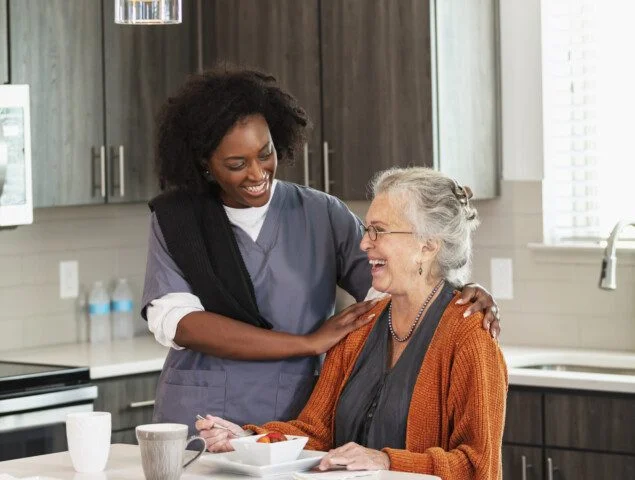 senior woman seated at the island in her home smiles while enjoying breakfast in the company of her caregiver
