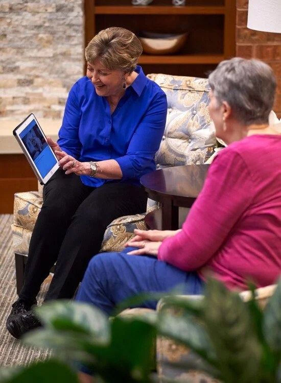 senior woman in blue shirt smiles as she shows her companion an image on her electronic tablet