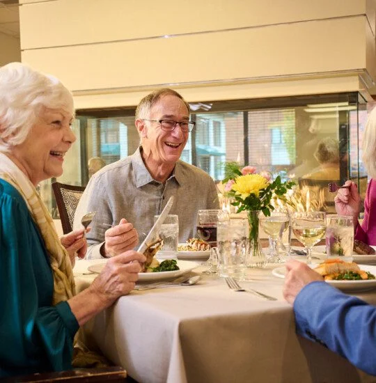 Four people share laughter and food at a dining table indoors.