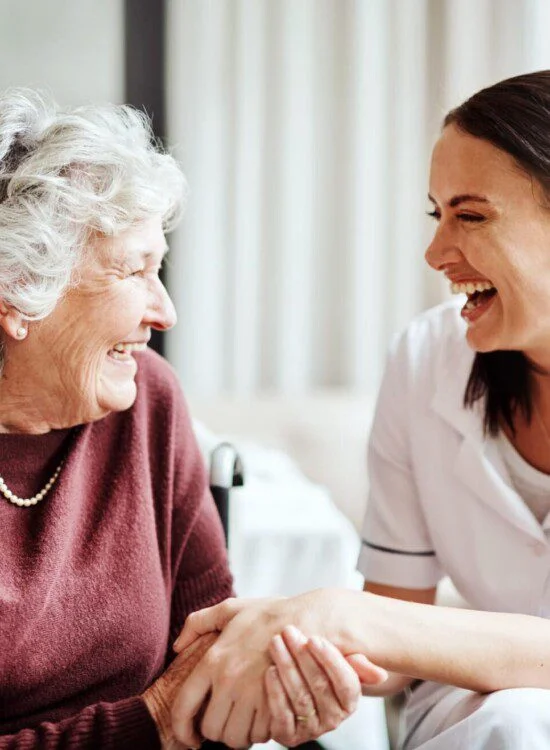 seated senior woman and her female caregiver laugh and hold hands while conversing