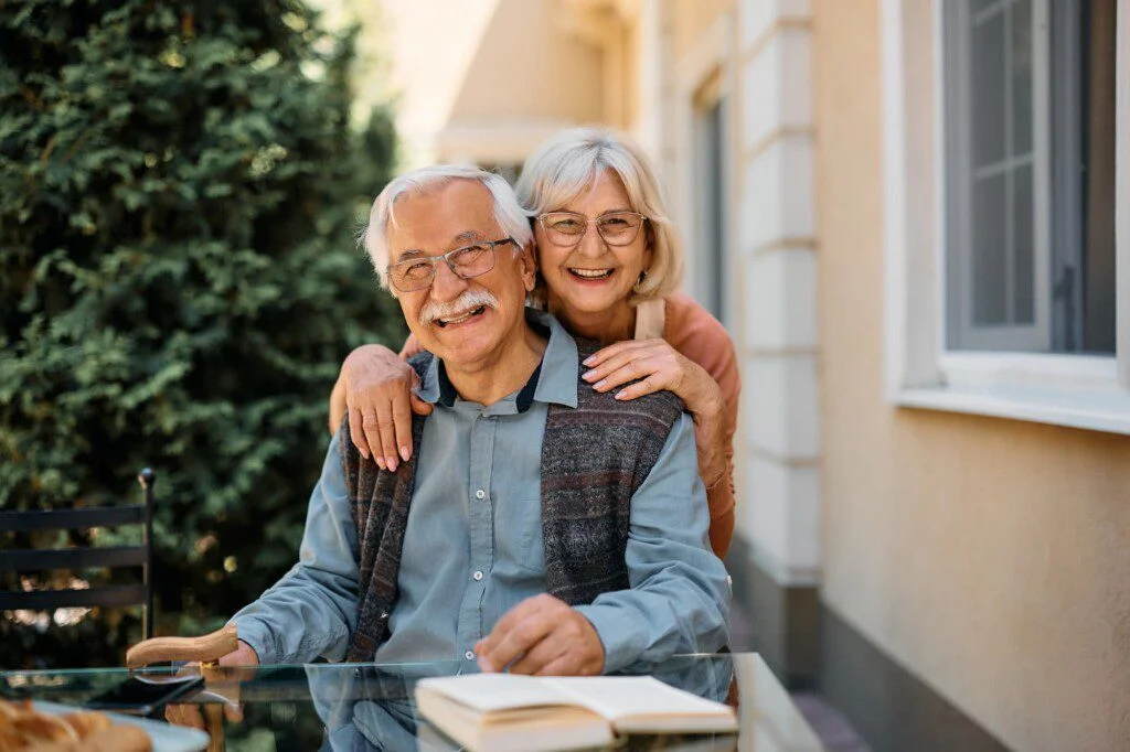 An elderly women hugging an elderly man from behind as he sits on a chair outside with a table in front