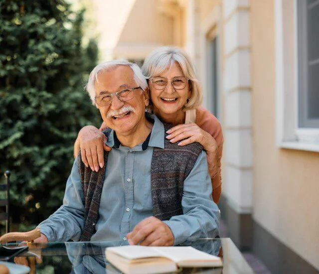 An elderly women hugging an elderly man from behind as he sits on a chair outside with a table in front