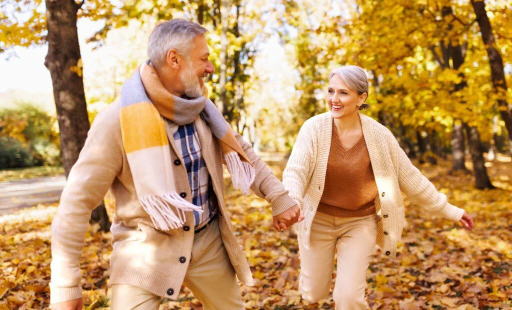 an elderly man and elderly woman holding hands and smiling at each other as they move through the woods during the fall season
