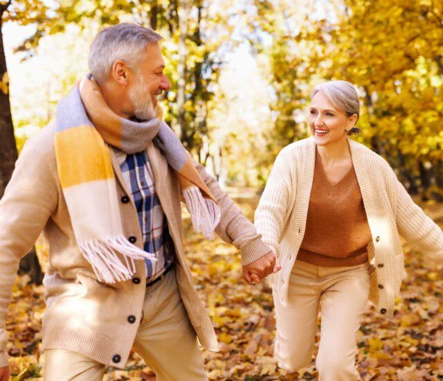 an elderly man and elderly woman holding hands and smiling at each other as they move through the woods during the fall season