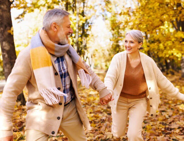 an elderly man and elderly woman holding hands and smiling at each other as they move through the woods during the fall season