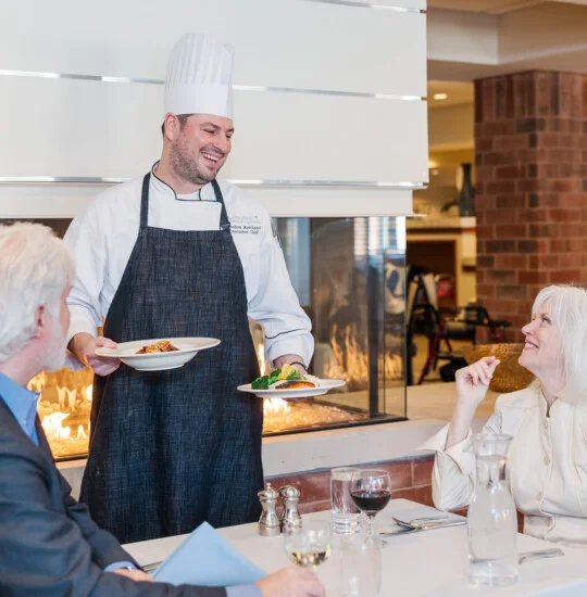 Chef serves dishes to couple at cozy fireplace restaurant setting.