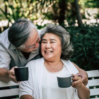 senior man stands behind seated wife to kiss her cheek, both holding coffee cups