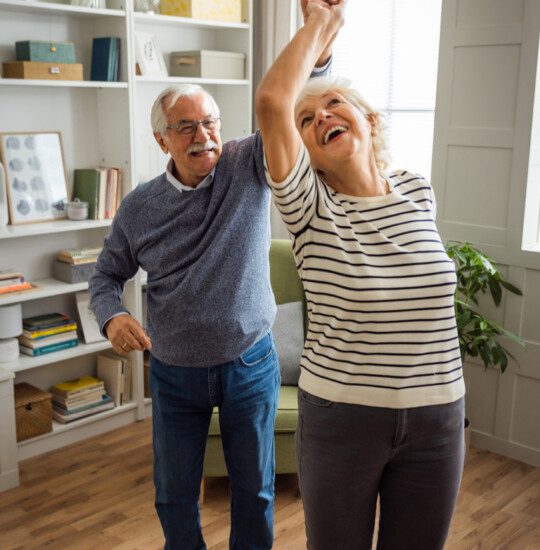 senior couple laugh and dance together in their apartment