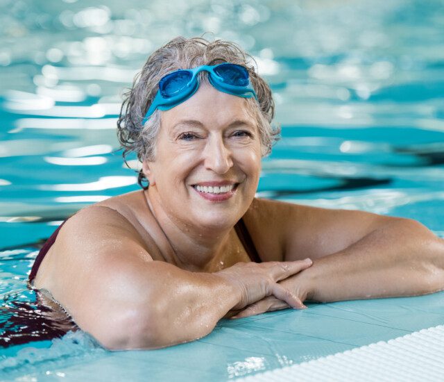 senior woman with swimming goggles on her head leans against the pool edge and smiles at the camera