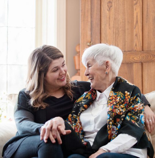 senior woman and adult daughter sit on a couch close together, smiling and holding hands