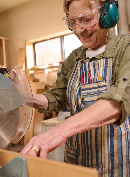 senior woman with protective glasses and headphones smiles while using saw to complete woodworking project
