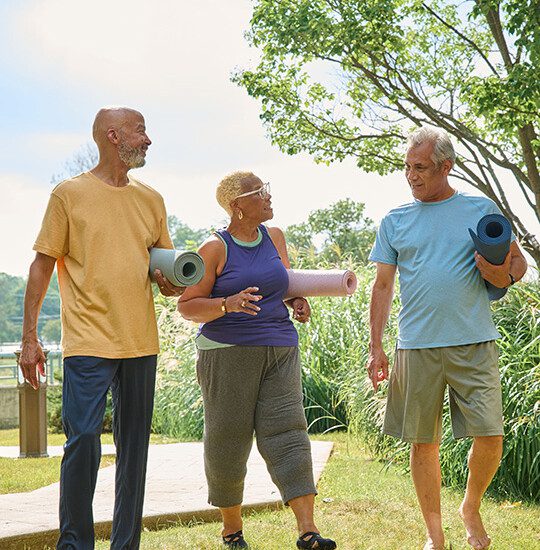 group of three seniors walk back to the path after a morning yoga session outside of their senior living community, backdropped by beautiful landscaping