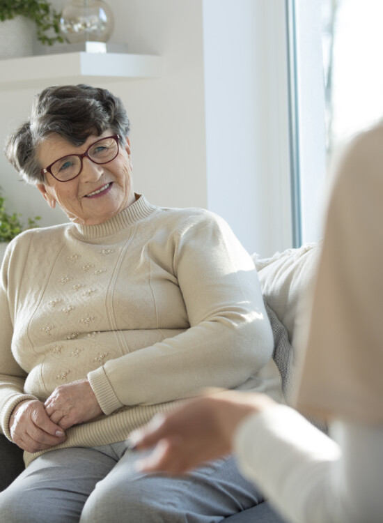 senior woman sitting on sofa talking to caregiver with their back to audience