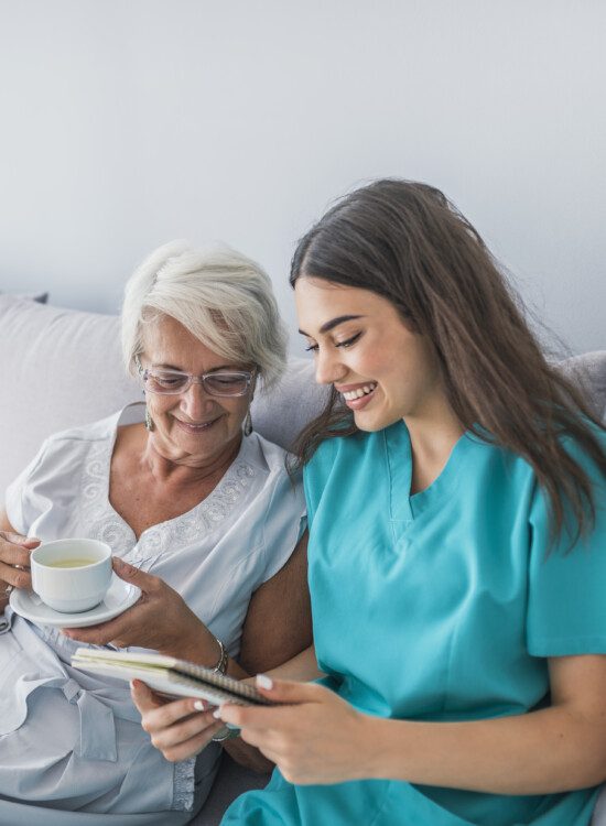 senior woman drinking coffee on her bed is shown information by her caretaker while seated