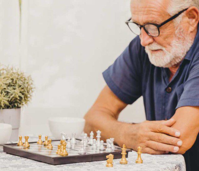 senior man in glasses and blue shirt sits at a table set with chess board