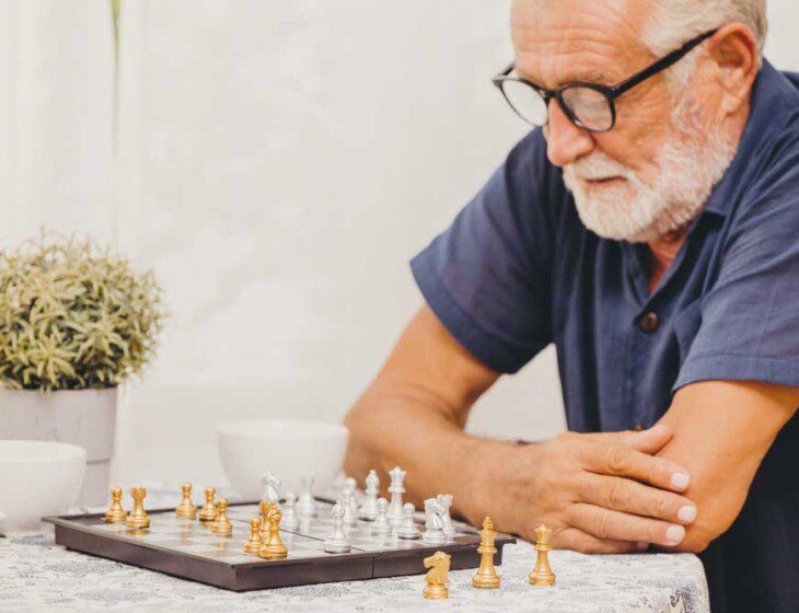 senior man in glasses and blue shirt sits at a table set with chess board