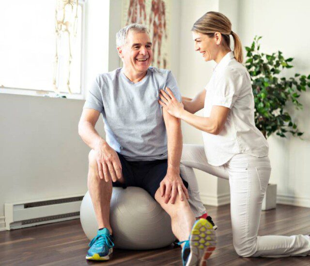 senior man on exercise ball extends his leg out and balances with the help of his physical therapy trainer