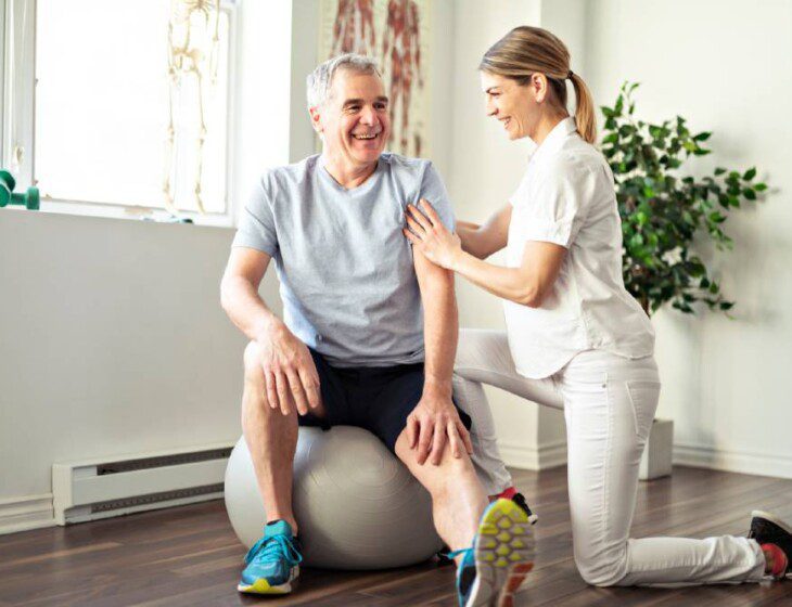 senior man on exercise ball extends his leg out and balances with the help of his physical therapy trainer