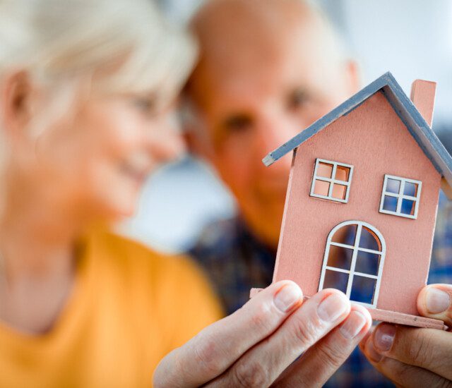 close-up of tiny home model being held by senior couple, blurred in the background