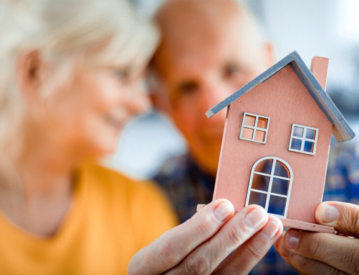 close-up of tiny home model being held by senior couple, blurred in the background