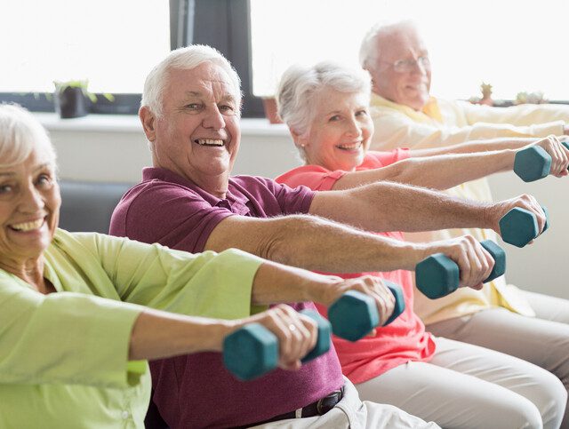 group of seniors raise dumbbells and smile at the camera during a seated fitness class