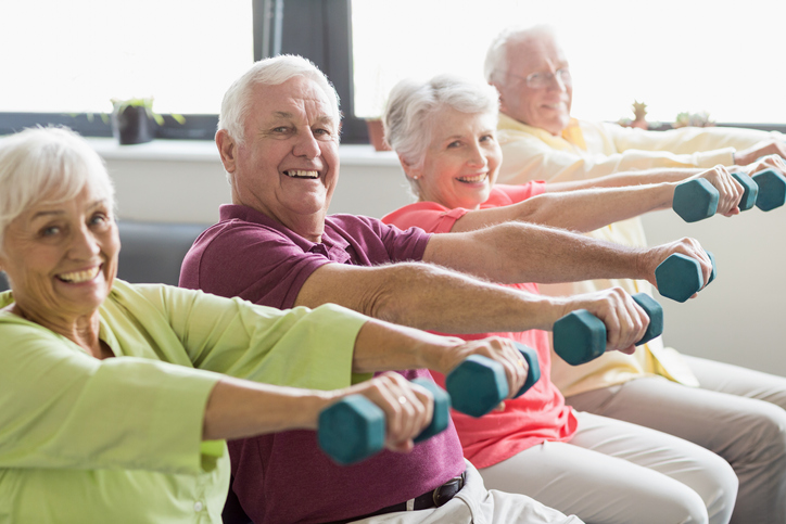 group of seniors raise dumbbells and smile at the camera during a seated fitness class