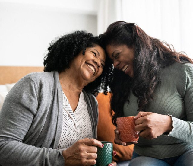 senior woman and her adult daughter sit on a bed, holding mugs of coffee and pressing their foreheads together, smiling