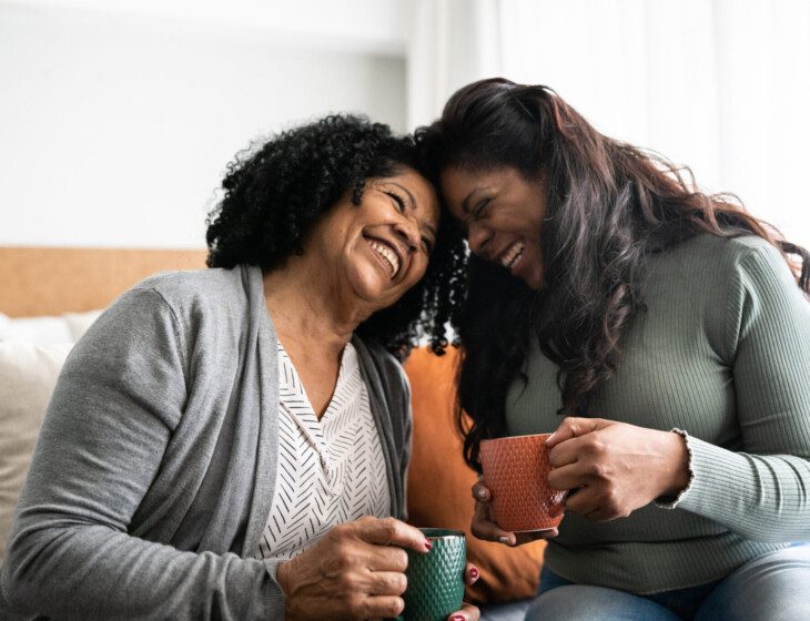 senior woman and her adult daughter sit on a bed, holding mugs of coffee and pressing their foreheads together, smiling