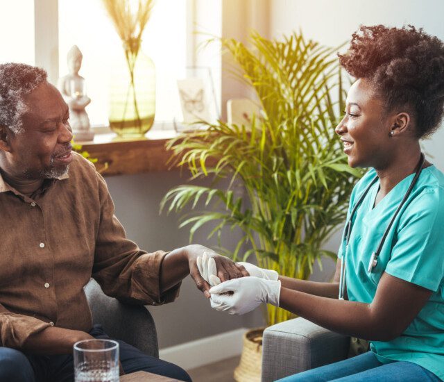 seated senior man smiles at his caregiver, who sits across from him holding his hand while wearing gloves