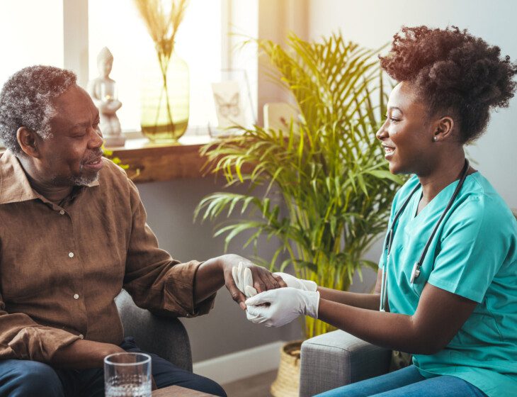 seated senior man smiles at his caregiver, who sits across from him holding his hand while wearing gloves