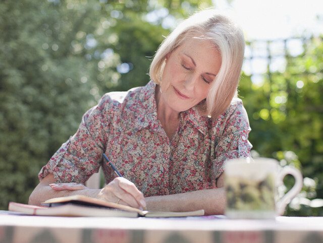 senior woman smiles softly while journaling at a table outdoors, backdropped by green trees