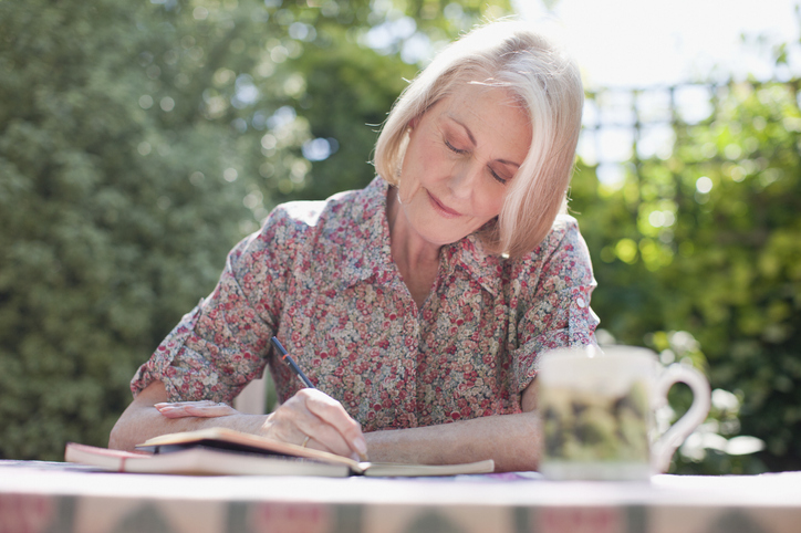 senior woman smiles softly while journaling at a table outdoors, backdropped by green trees