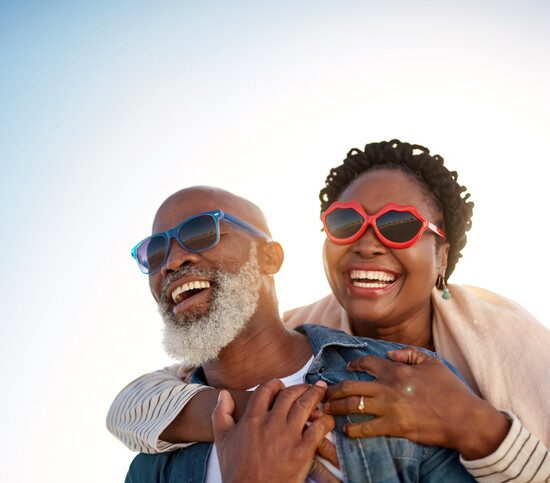 senior woman receives piggy back ride from her husband, both wearing sunglasses
