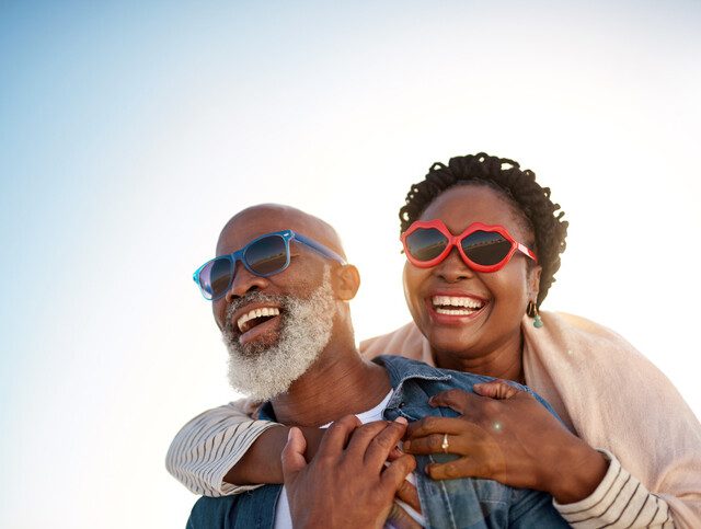 senior woman receives piggy back ride from her husband, both wearing sunglasses