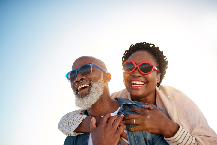 senior woman receives piggy back ride from her husband, both wearing sunglasses