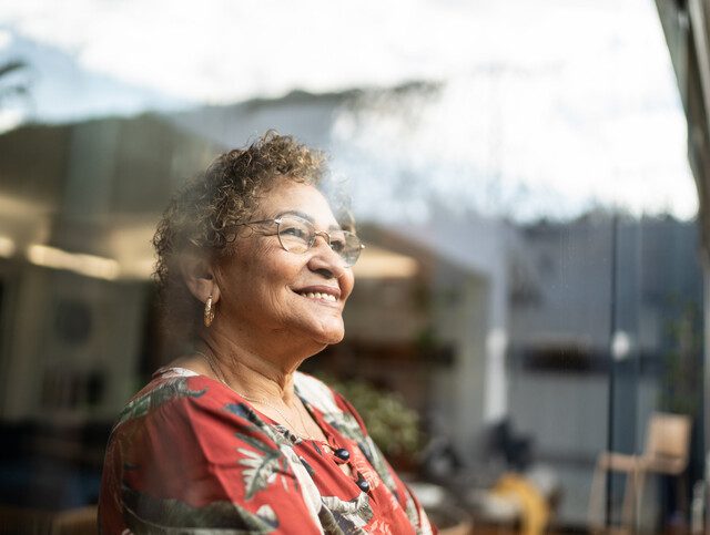 senior woman in glasses and patterned blouse smiles while looking out of a window