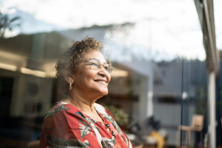 senior woman in glasses and patterned blouse smiles while looking out of a window