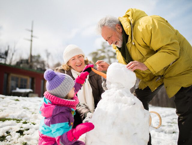 senior couple in winter coats smile while helping their granddaughter build a snowman