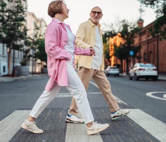 stylish senior man and woman hold hands while crossing the street