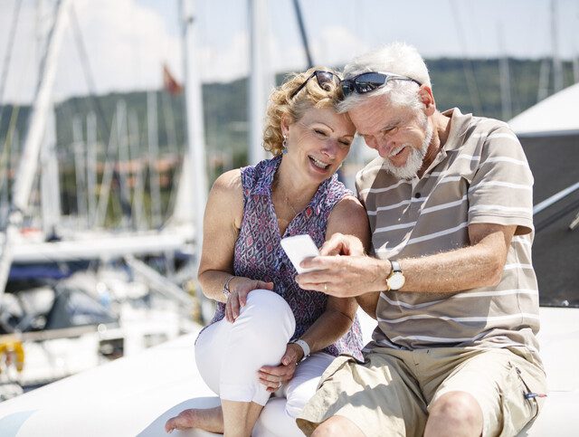 stylish senior couple smile at their phone while sitting on a small yacht in the harbor