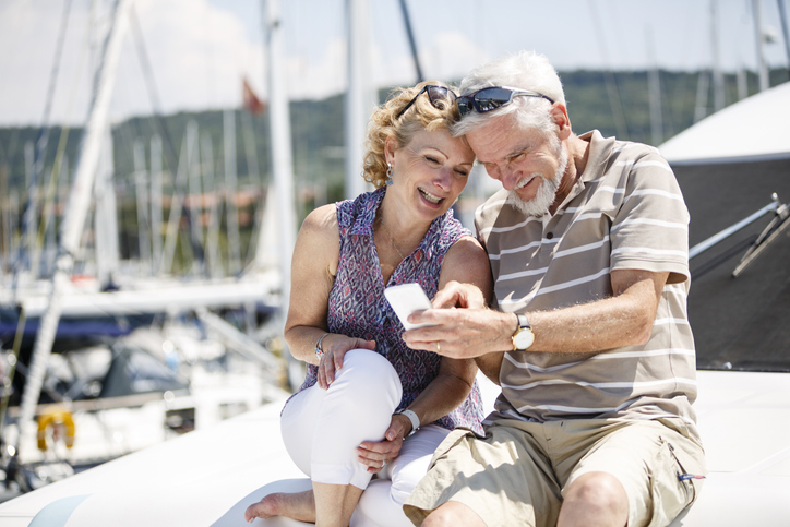 stylish senior couple smile at their phone while sitting on a small yacht in the harbor