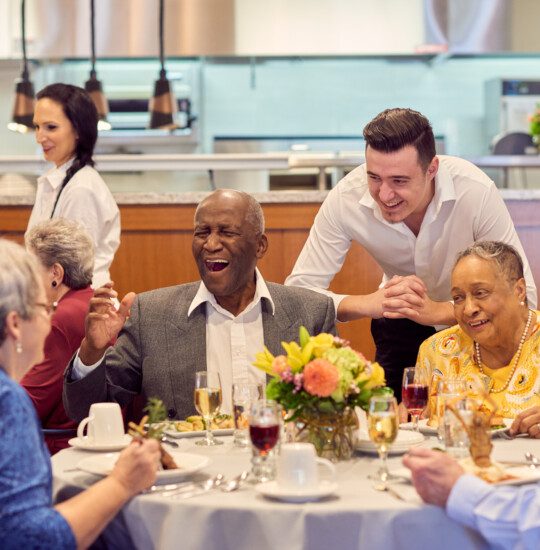 two tables of seniors laugh and converse while enjoying dinner and wine together inside