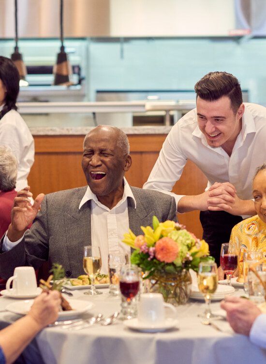 two tables of seniors laugh and converse while enjoying dinner and wine together inside