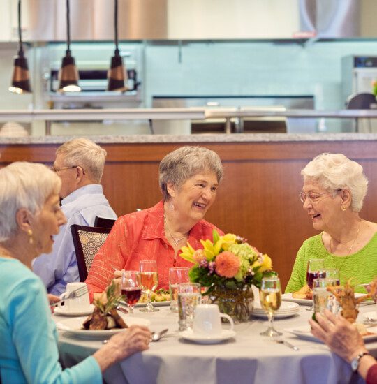 two tables of seniors laugh and converse while enjoying dinner and wine together inside