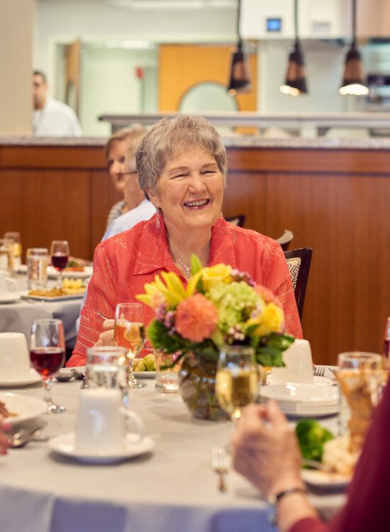 two tables of seniors laugh and converse while enjoying dinner and wine together inside