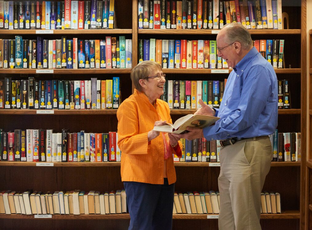 senior man and woman smile and discuss a book held by the man while in the library of their senior living community